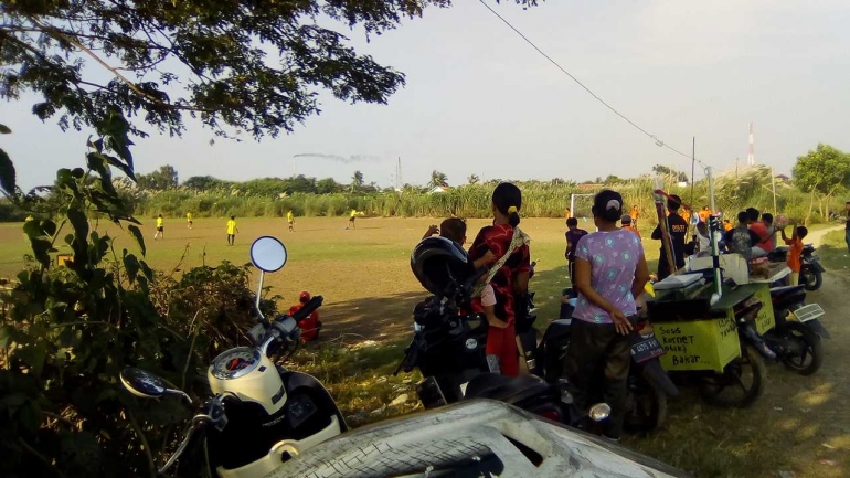 beberapa warga desa sedang menyaksikan pertandingan sepakbola kampung, beberapa pedagang juga sudah siap menjajakan dagangannya