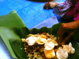 nasi kuning yang berisi telur dadar, tempe orek, perkedel dan kerupuk dinikmati dengan tangan tanpa sendok makan