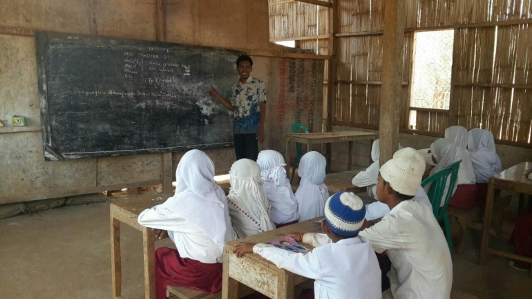 Guru sedang mengajar di kelas yang dindingnya hanya terbuat dari bambu dengan alas tanah | Sumber: kitabisa.com/motoruntukpakguru