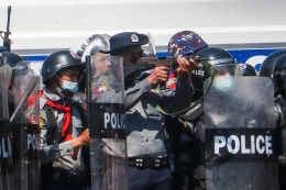 Seorang polisi (tengah) mengacungkan senapannya dalam bentrokan melawan massa yang ikut dalam demonstrasi menentang kudeta militer Myanmar di Naypyidaw, pada 9 Februari 2021. (STR via AFP/kompas.com)