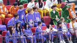 Sumber: Soompi. TWICE dan Stray Kids yang memakai jaket yang sama sedang duduk di kursi penonton sambil menyemangati anggota mereka masing - masing