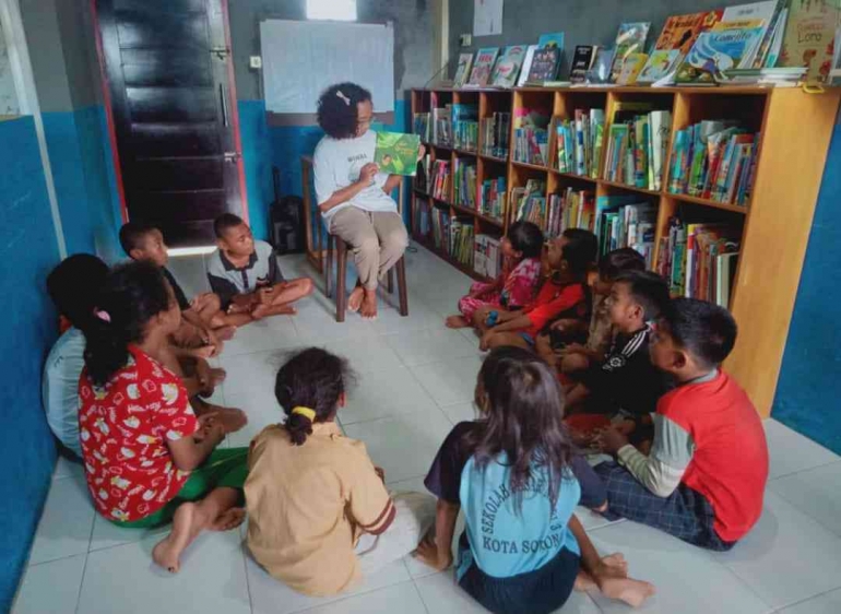 Gambar 2. Grace Burdam, salah seorang relawan pustakawan sedang membaca nyaring bagi pengunjung yang datang. (Sumber foto : pribadi)