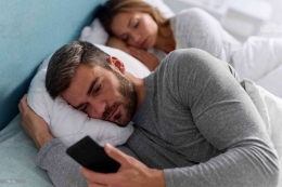 Sumber. Pemuda menggunakan ponsel dan memeriksa pesan di tempat tidur sementara istrinya sedang tidur/istock.