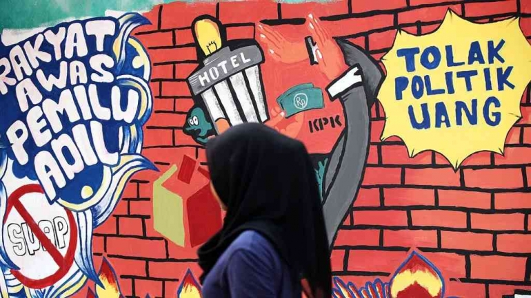 Seorang perempuan melintas di depan mural anti politik uang. Sumber : Kompas.id