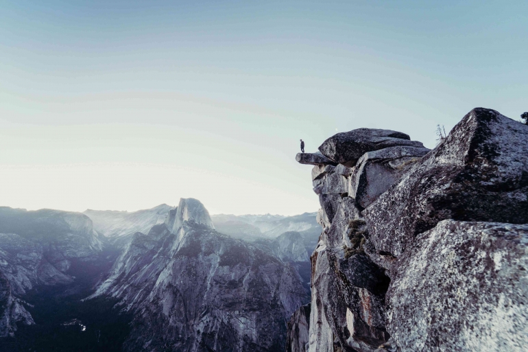 Ilustrasi: Seorang laki-laki yang berdiri di tepi gunung batu yang spektakuler di Yosemite National Park. Sumber: Unsplash / Cristofer Maximilian